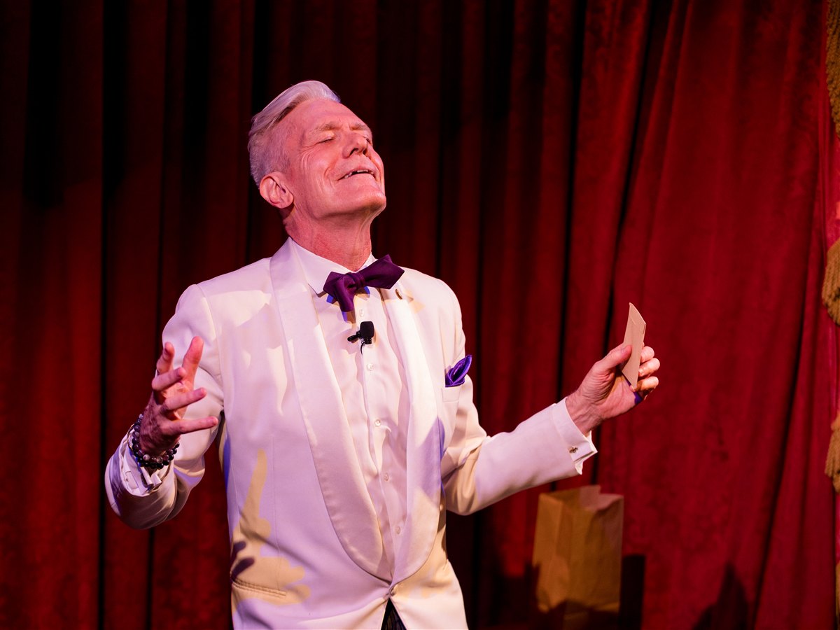 Felix Jones mid-performance with arms raised, holding a card, on stage at the Magic Castle